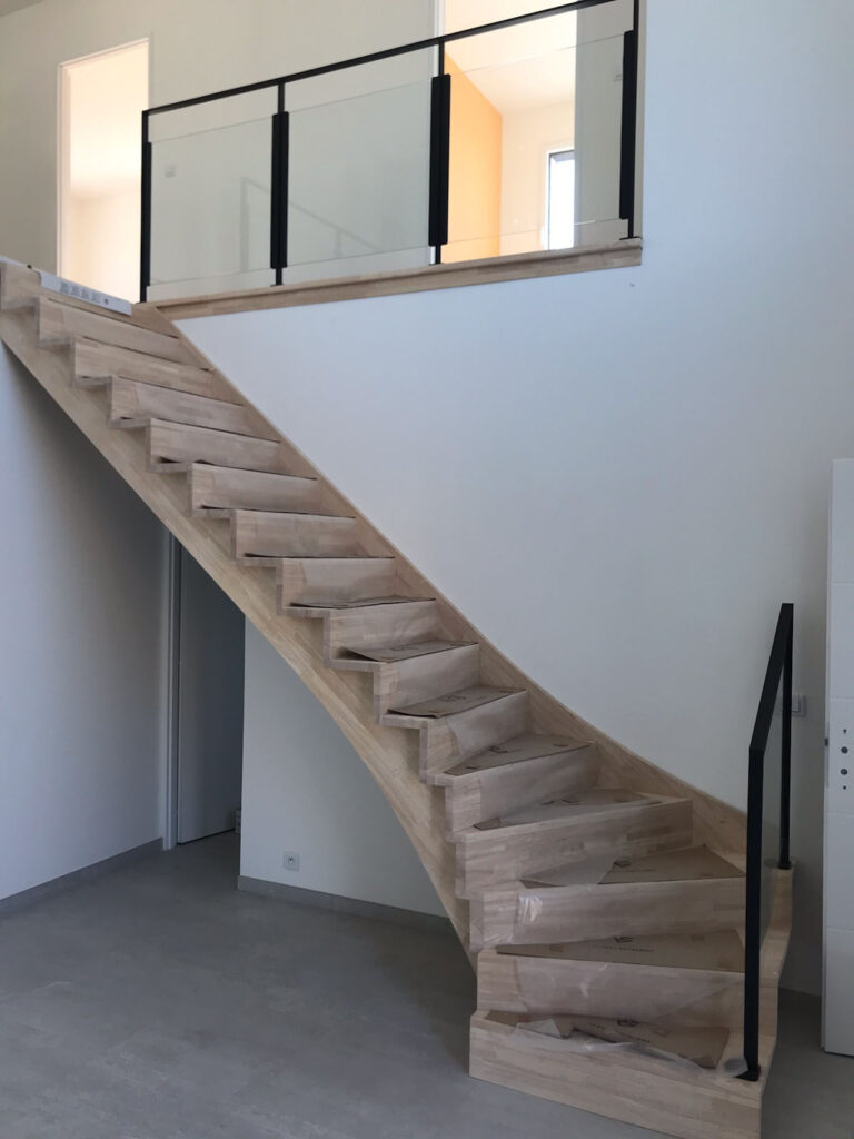 Escalier quart-tournant en bois clair menant à une mezzanine avec garde-corps vitré noir.